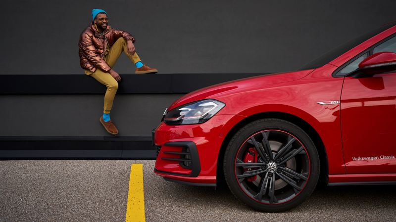 A man sits on a ledge, in the foreground the front of a red VW in side view