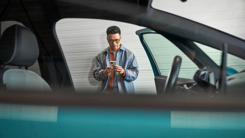 Blick durch die offene Fahrertür eines VW ID. Modells auf einen Mann mit Smartphone, der an einer Wand lehnt