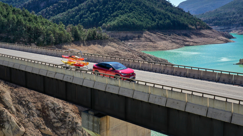 Ένα VW ID.5 GTX που μεταφέρει με τρέιλερ ένα καγιάκ οδηγεί πάνω σε μια γέφυρα με μια λίμνη στο βάθος – λύσεις μεταφοράς από τα Αξεσουάρ VW