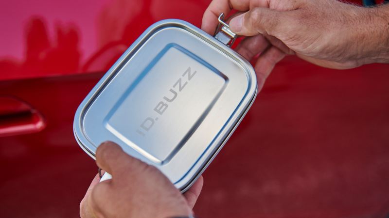 Jemand hält einen Essensbehälter aus Edelstahl mit „ID. Buzz“-Gravur in den Händen – VW Zubehör