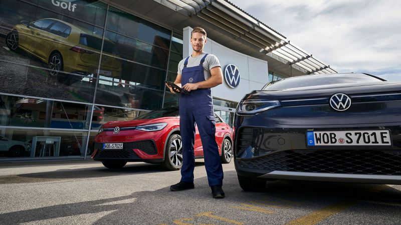 A VW service employee with a tablet in his hand in front of two VW cars next to a workshop