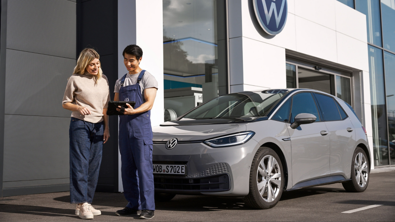 A VW service employee with tablet and a customer in front of VW car next to a workshop