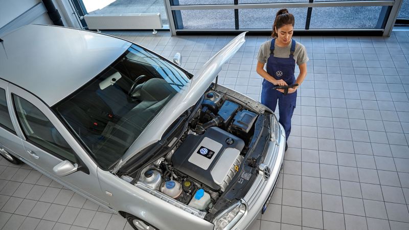 VW Servicemitarbeiterin neben einem VW Golf 4 mit offener Motorhaube schaut auf ein Tablet