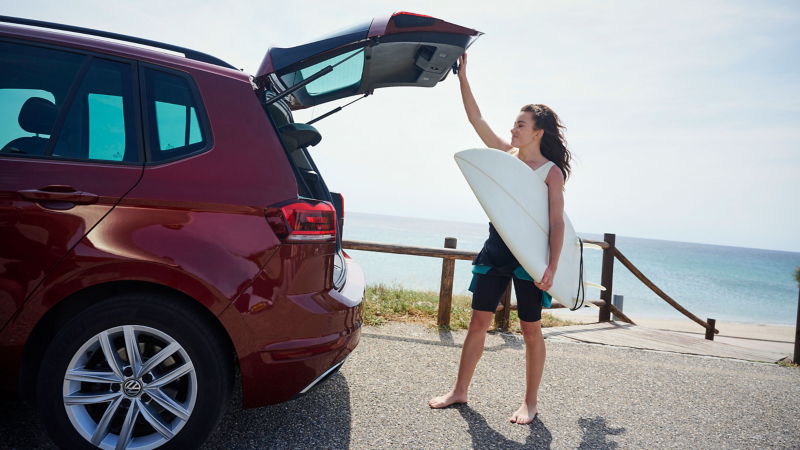 Une femme avec une planche de surf se tient devant le coffre ouvert d’une VW Golf Sportsvan – accessoires pour les anciens modèles