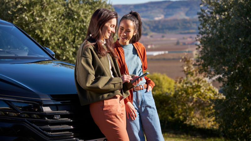 Two women in nature leaning against a VW Touareg R model and looking at a smartphone