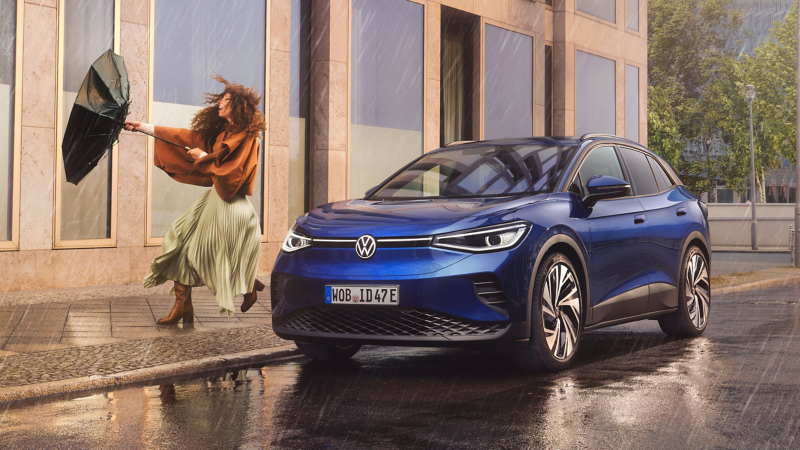 Frau im Regen hält wegfliegenden Schirm fest, daneben VW ID.4 Pro in Blau am Straßenrand