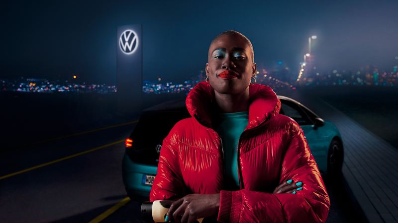Une femme vêtue d’une veste rouge se tient devant une VW ID. et un logo VW illuminé
