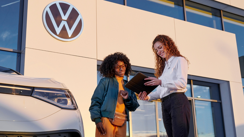 VW Servicemitarbeiterin und Kundin schauen auf ein Tablet, daneben ein VW ID.5
