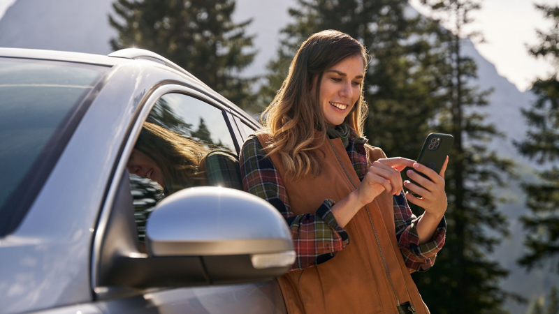 Eine Frau lehnt an einem VW Auto und tippt etwas auf ihrem Smartphone ein