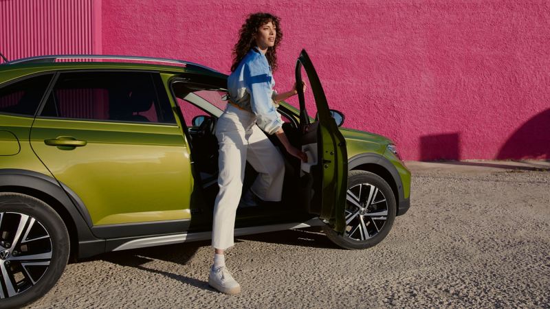 Una chica bajando en un Volkswagen Taigo verde frente a un muro color frambuesa