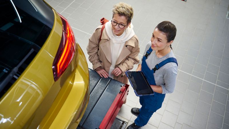 VW Golf sur une plate-forme élévatrice durant le contrôle en atelier préalable au contrôle technique officiel, à côté d'un client et d’un collaborateur de service VW