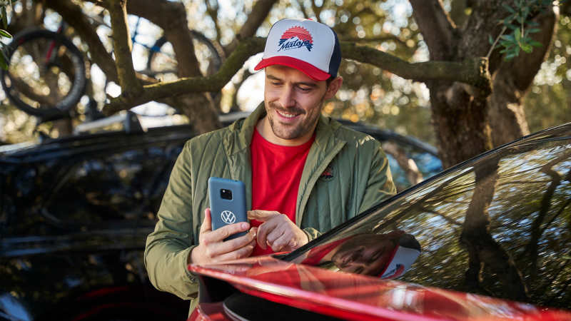 Mann steht neben VW Auto und schaut auf ein Smartphone