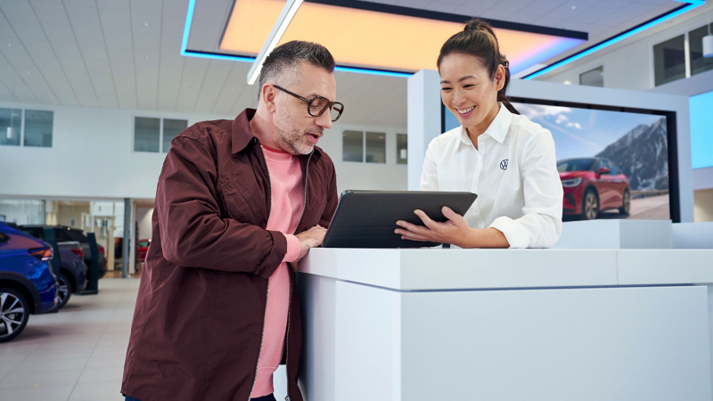 Kunde und VW Servicemitarbeiterin an einem Pult im Autohaus schauen gemeinsam auf ein Tablet
