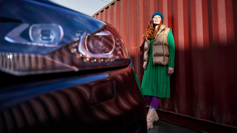 Close-up headlight of a VW Touareg 2, woman leaning against a red wall next to the car and enjoying the sun