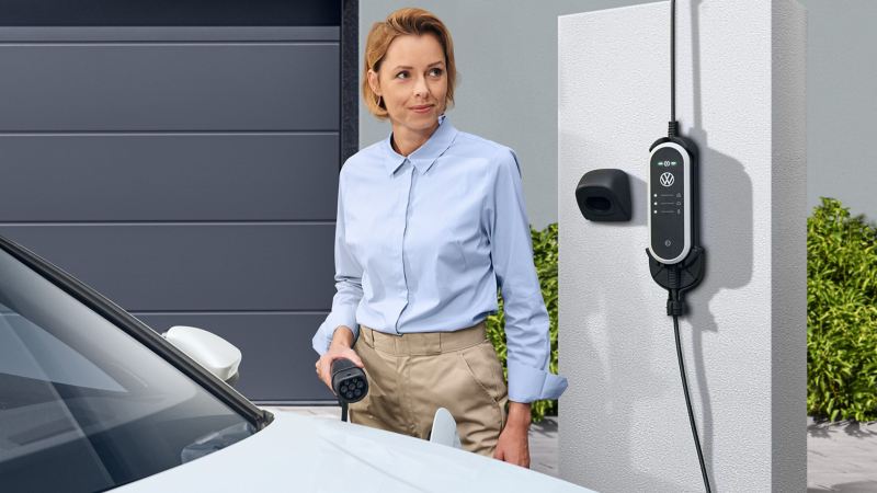 Woman with charging cable in hand in front of a VW Passat model, wall mount set ID. Charger Travel Basic in the background