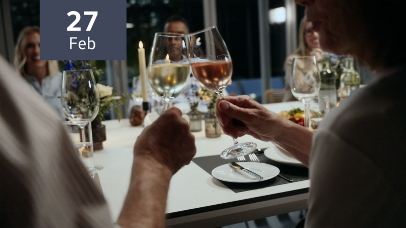 Gäste sitzen an festlich geschmückter Tafel, zwei Personen prosten sich mit Weingläsern zu