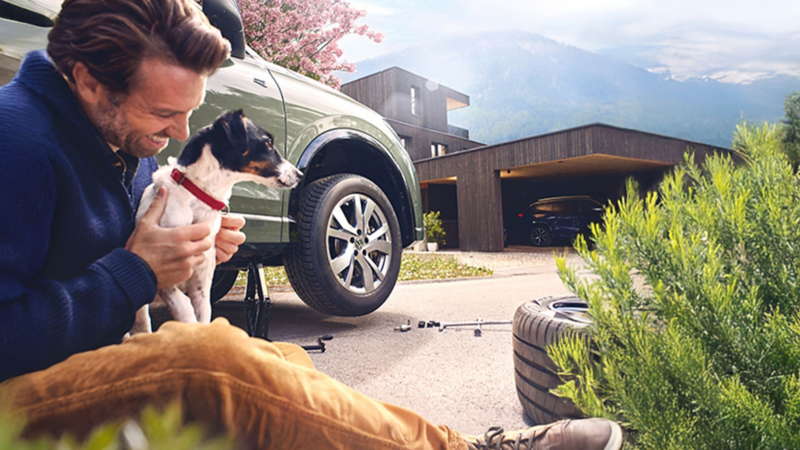 Mann mit Hund sitzt vor VW Auto beim Reifenwechsel