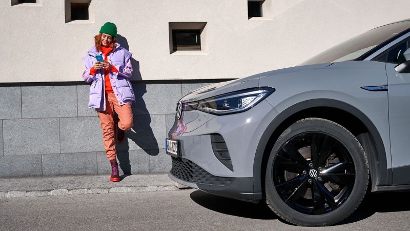 Kundin steht neben VW Elektroauto und informiert sich am Smartphone zur Reifengarantie