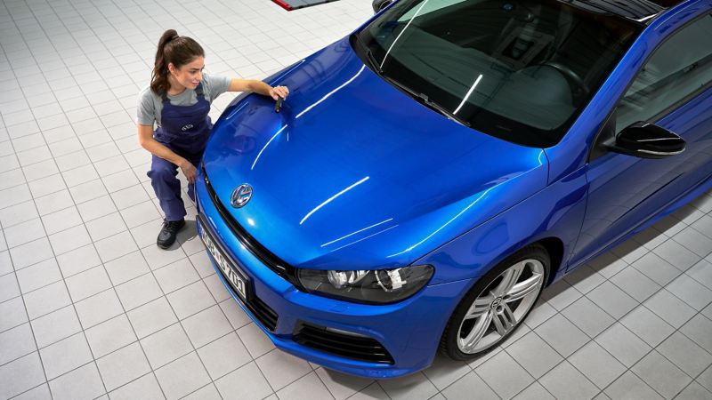 VW Servicemitarbeiterin untersucht Lack von Motorhaube eines VW Scirocco 3