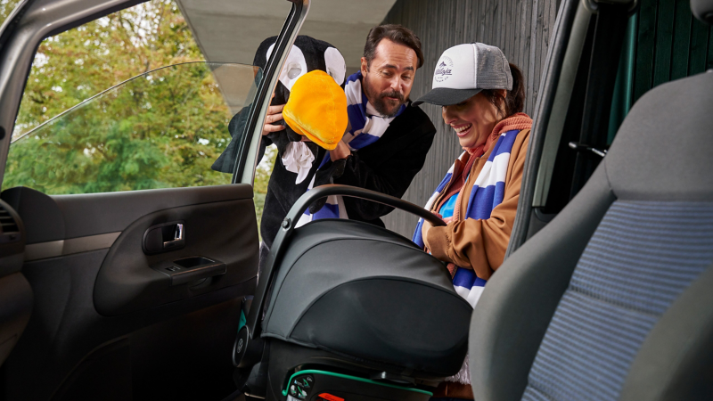 Une femme retire le siège bébé d’une voiture VW, un homme amuse l’enfant avec une peluche – accessoires pour les modèles précédents