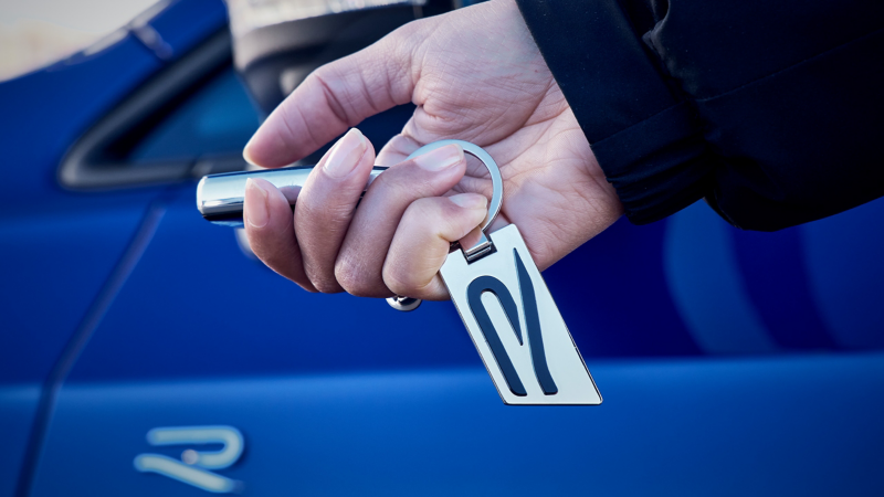 Silberner Schlüsselring mit schwarzem R-Logo, der an einem Autoschlüssel befestigt ist, im Hintergrund blaues VW R-Modell