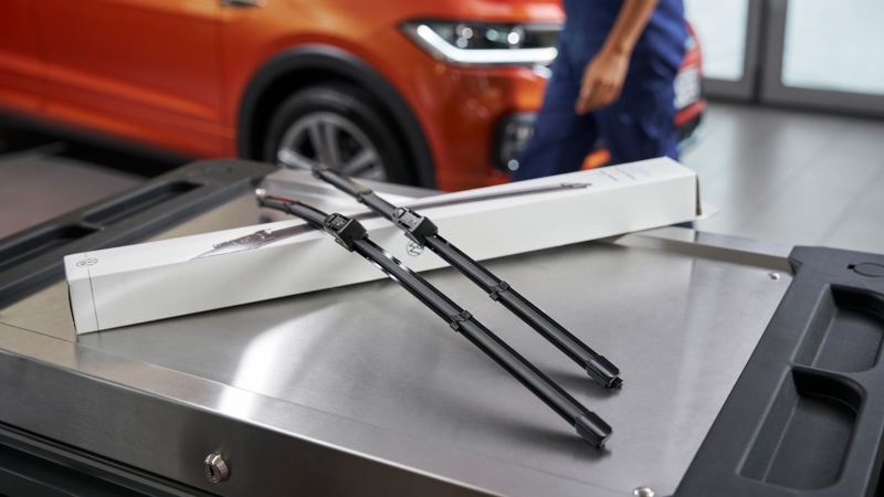 Essuie-glaces Aero d’origine Volkswagen à côté de leur emballage sur un conteneur roulant, un Volkswagen T-Cross en arrière-plan