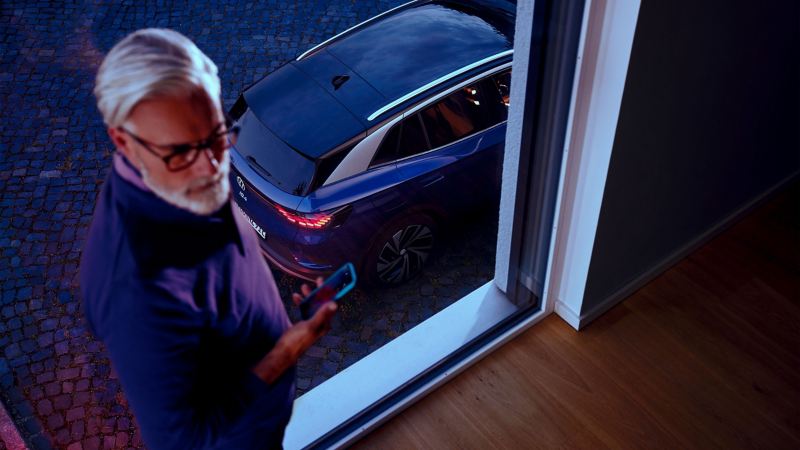 Ein Mann steht am Fenster mit seinem Smartphone in der Hand, sein ID. parkt draußen