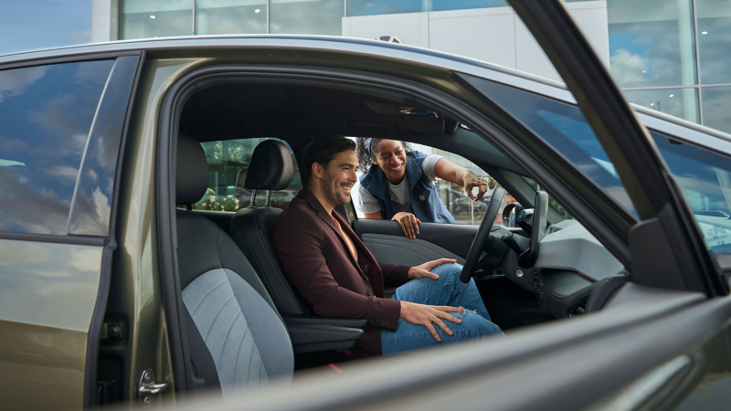Mężczyzna siedzi na fotelu kierowcy w Volkswagenie. Przez otwarte okno zagląda pracownica serwisu VW i wskazuje palcem kokpit.