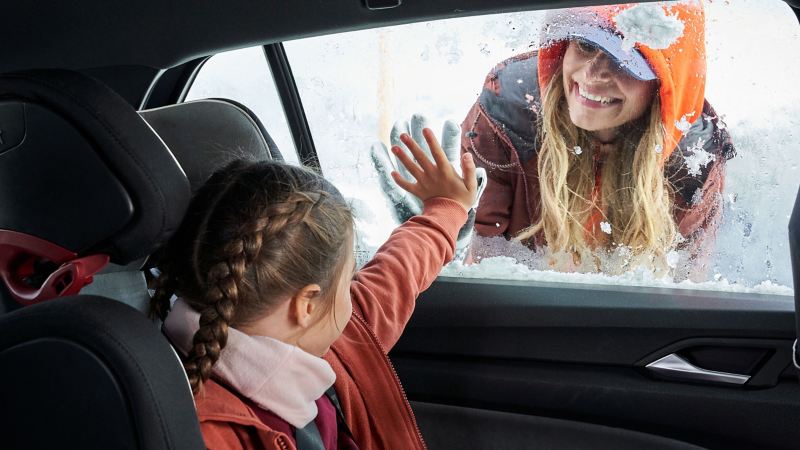 Eine Mutter schaut durch die zugefrorene Scheibe in den warmen Innenraum des Autos zu ihrer Tochter – Standheizung von VW