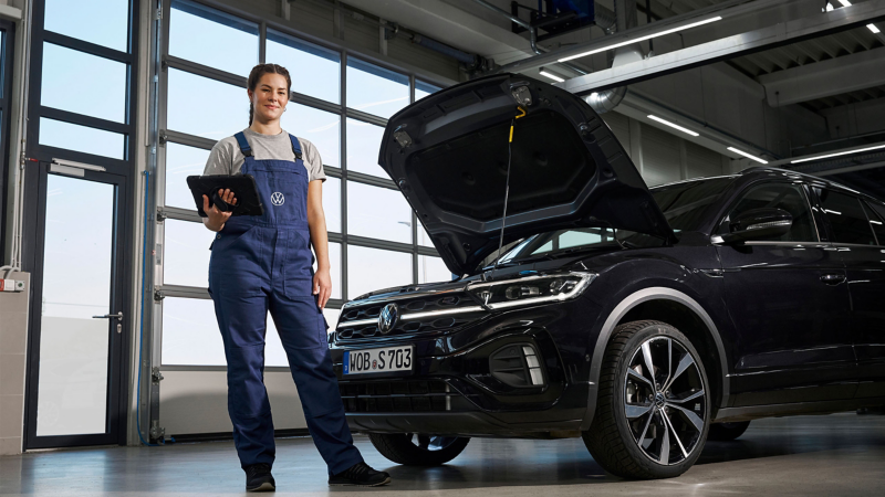 VW Servicemitarbeiterin neben VW T-Roc 1 mit geöffneter Motorraumklappe in Werkstatt