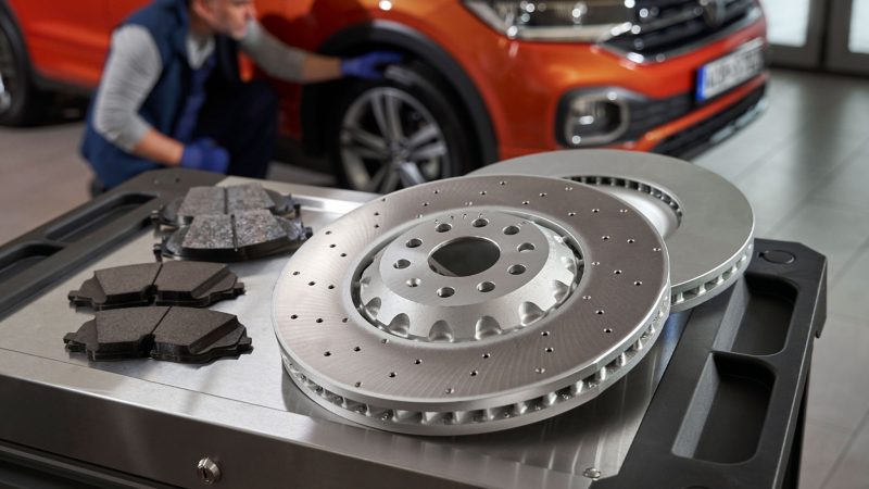 Volkswagen Original Bremsscheiben und Bremsbeläge auf einem Rollcontainer, im Hintergrund ein VW T-Cross – Bremsbeläge Auto