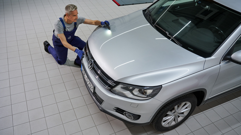 Service employee kneels in front of a VW Tiguan 1 and checks paintwork for damage