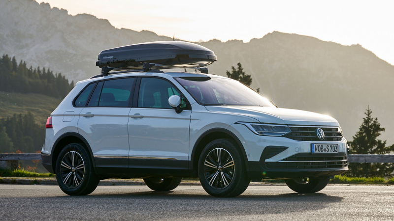 VW Tiguan mit Dachbox von VW Zubehör auf einer Landstraße, im Hintergrund Berge