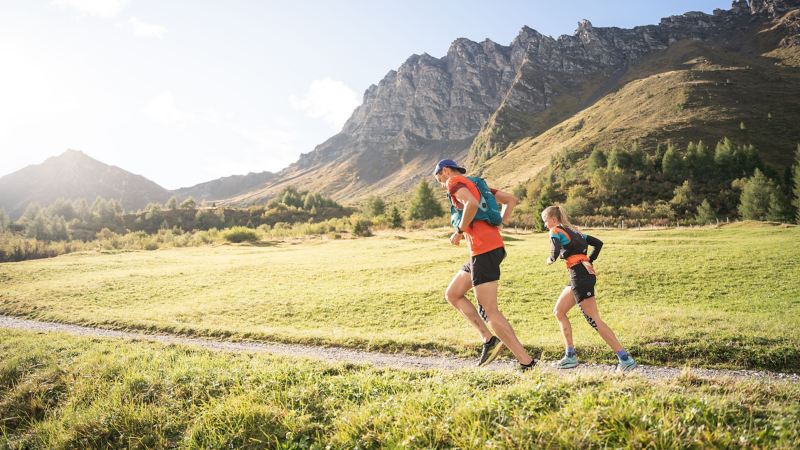 Ein Läuferteam läuft auf einem Feldweg, im Hintergrund Berge – Transalpine Run powered by VW R