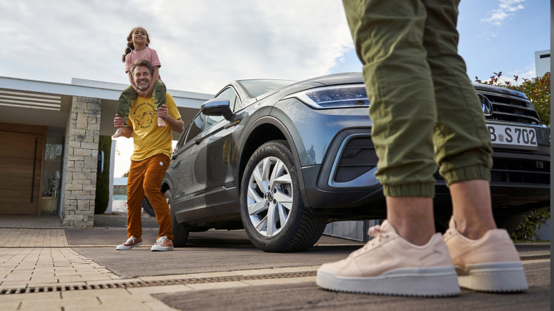 Family with Volkswagen in driveway, man with child on shoulders.