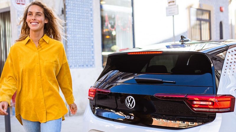 Eine Frau in einer gelben Bluse geht lächelnd von ihrem VW ID.3 weg