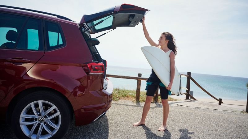 Eine Frau mit Surfbrett steht an der offenen Kofferraumklappe ihres VW Golf Sportsvan – Zubehör für Ihr Auto