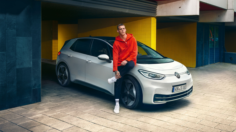 Un hombre con una chaqueta roja se apoya en su VW ID.