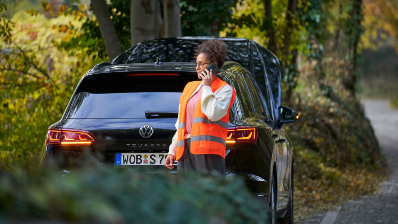 Frau mit Warnweste vor VW Touareg mit offener Motorhaube telefoniert mit Volkswagen Notdienst