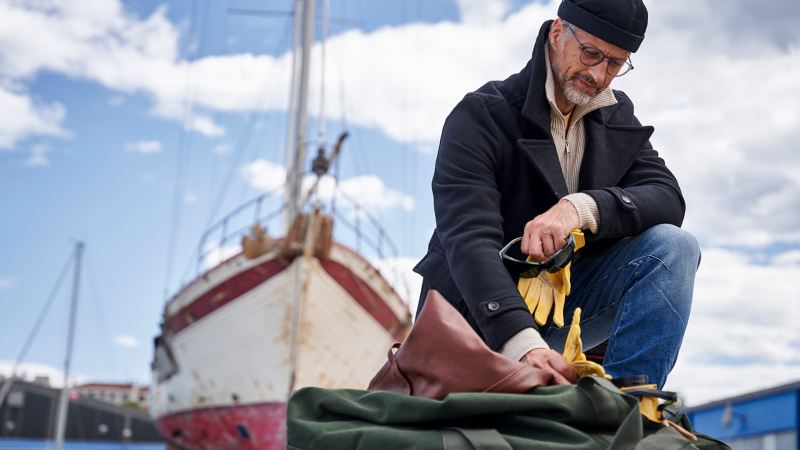 Der Hobbyhandwerker verstaut seine Arbeitskleidung in einer Tasche, im Hintergrund steht sein Segelboot