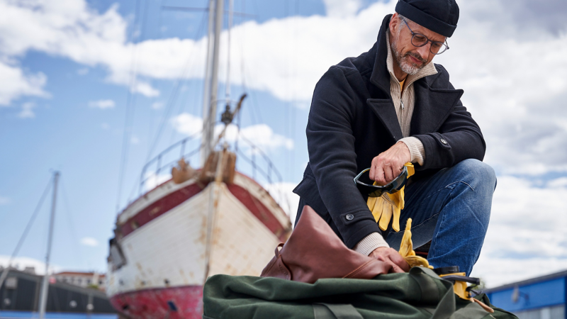 Hobbyhandwerker verstaut Arbeitskleidung in einer Tasche, im Hintergrund steht Segelboot