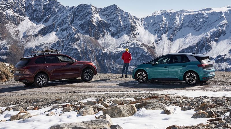 雪に覆われた山の景色を楽しんでいる男性の隣に2台のフォルクスワーゲン車――ウィンタータイヤ