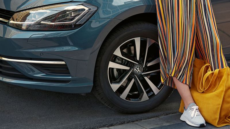 Una mujer con un bolso se apoya en su coche Volkswagen azul con una combinación llanta/neumático
