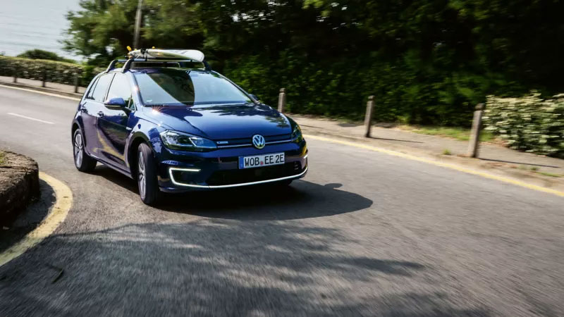 Une Volkswagen Golf électrique bleue roule sur une route sinueuse, avec un coffre de toit visible.