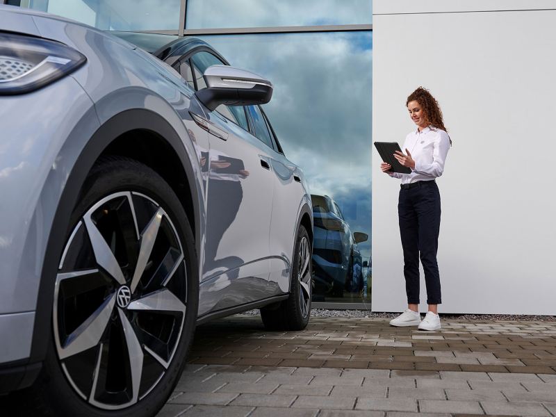 Mitarbeiterin aus dem VW Service steht mit einem Tablet neben einem VW Modell