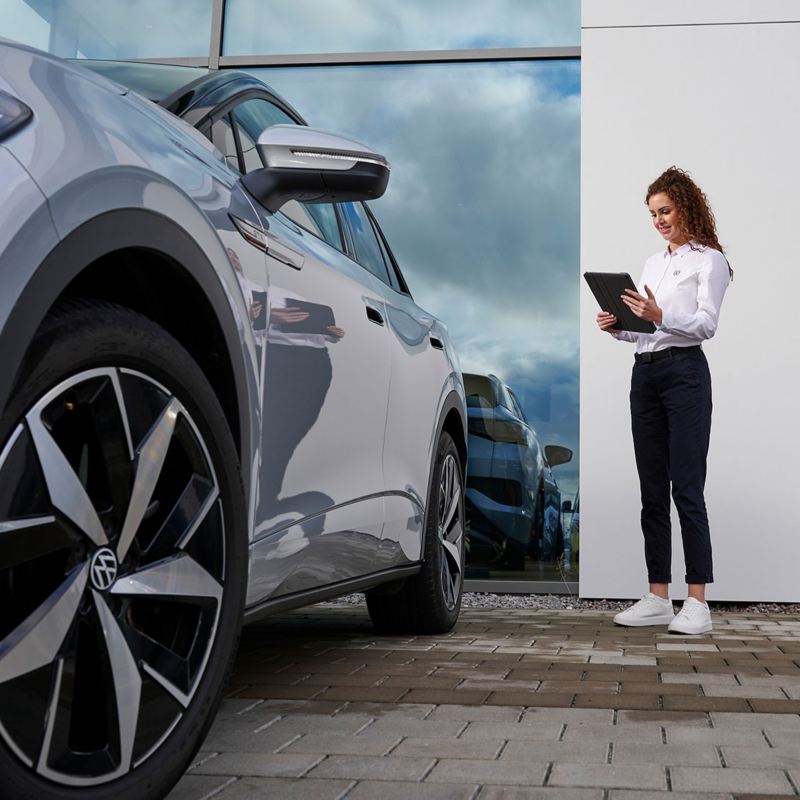 Mitarbeiterin aus dem VW Service steht mit einem Tablet neben einem VW Modell