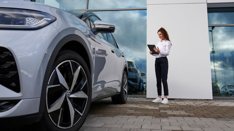 Mitarbeiterin aus dem VW Service steht mit einem Tablet neben einem VW Modell