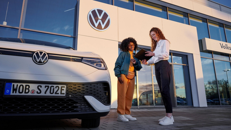 Eine Servicemitarbeiterin spricht mit einer Frau vor einem VW Händlergebäude, davor steht ein VW ID.5.