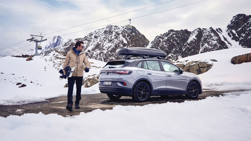 En mann med snowboard går vekk fra en vw id4 med takboks i fjellet
