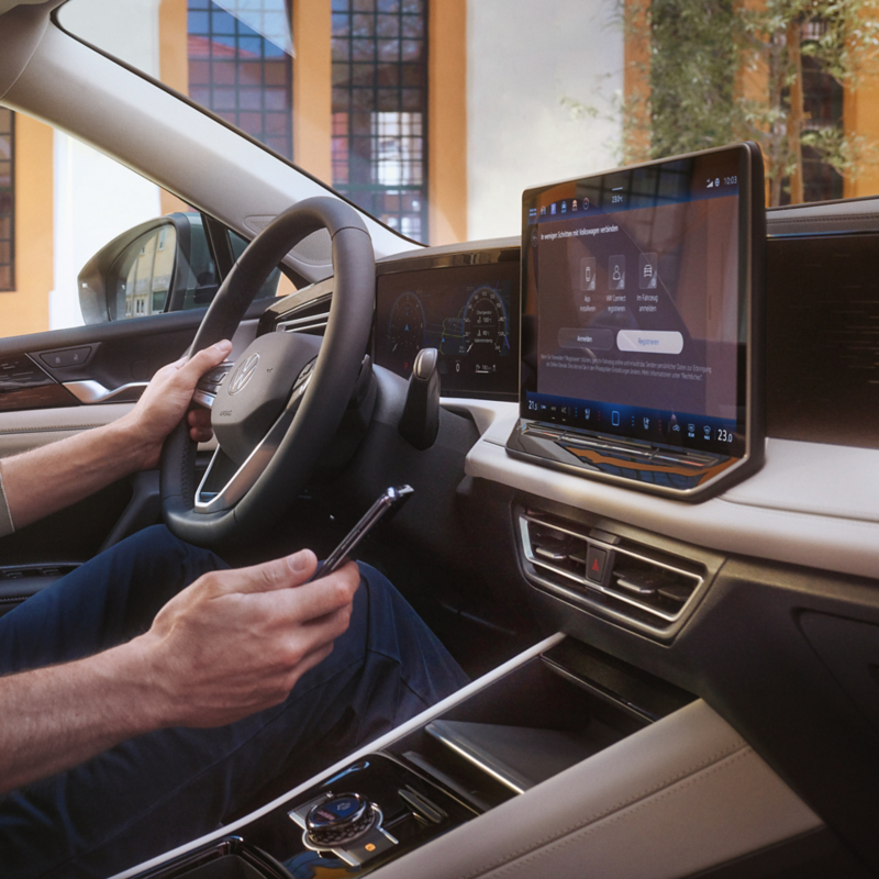 Innenschuss eines VW Tiguan. Ein Mann auf dem Fahrersitz steuert mit seinem Smartphone die optionale VW Connect Plus Anwendung.